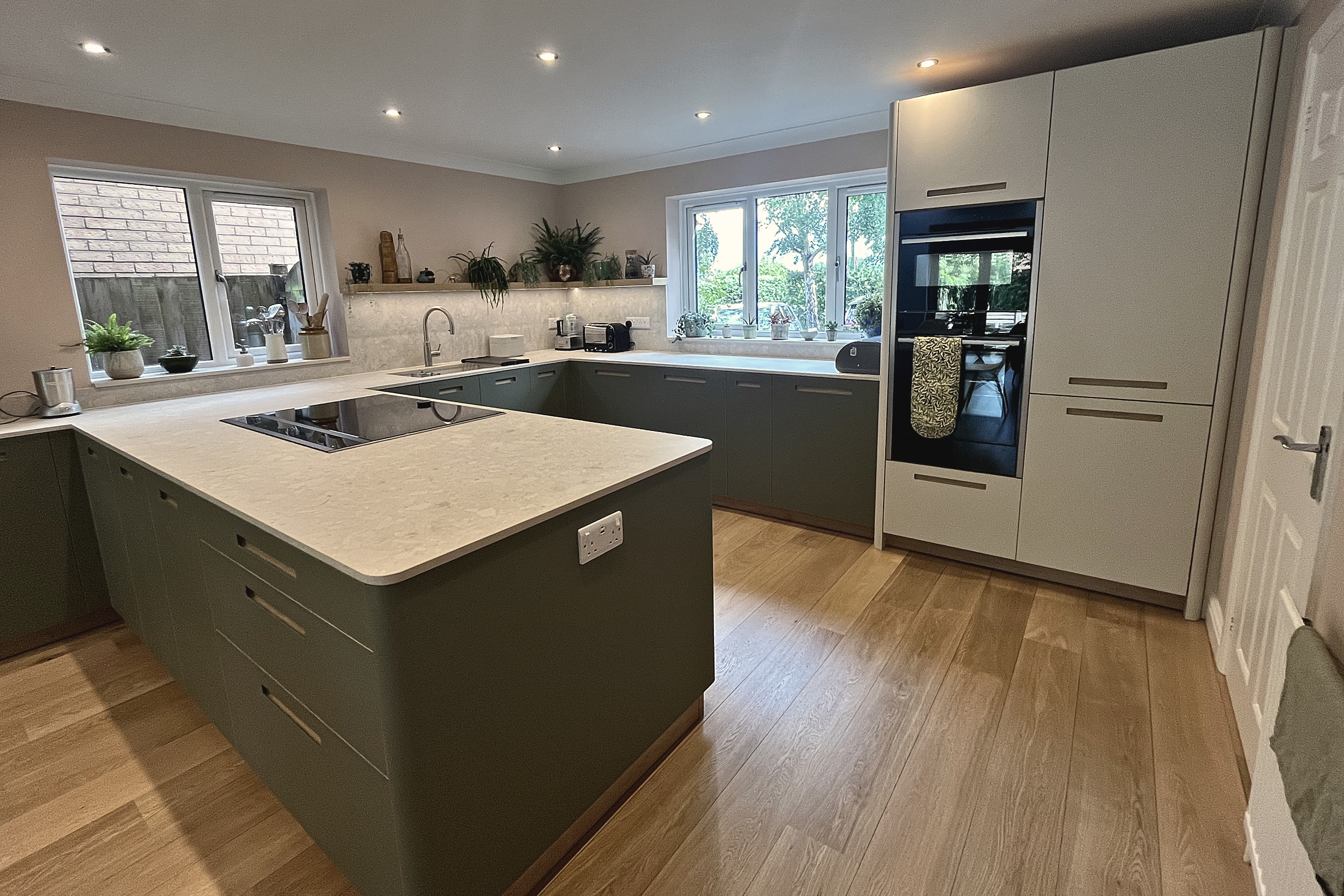 Green kitchen peninsula with induction hob, pale quartz worktop and tall oven housing