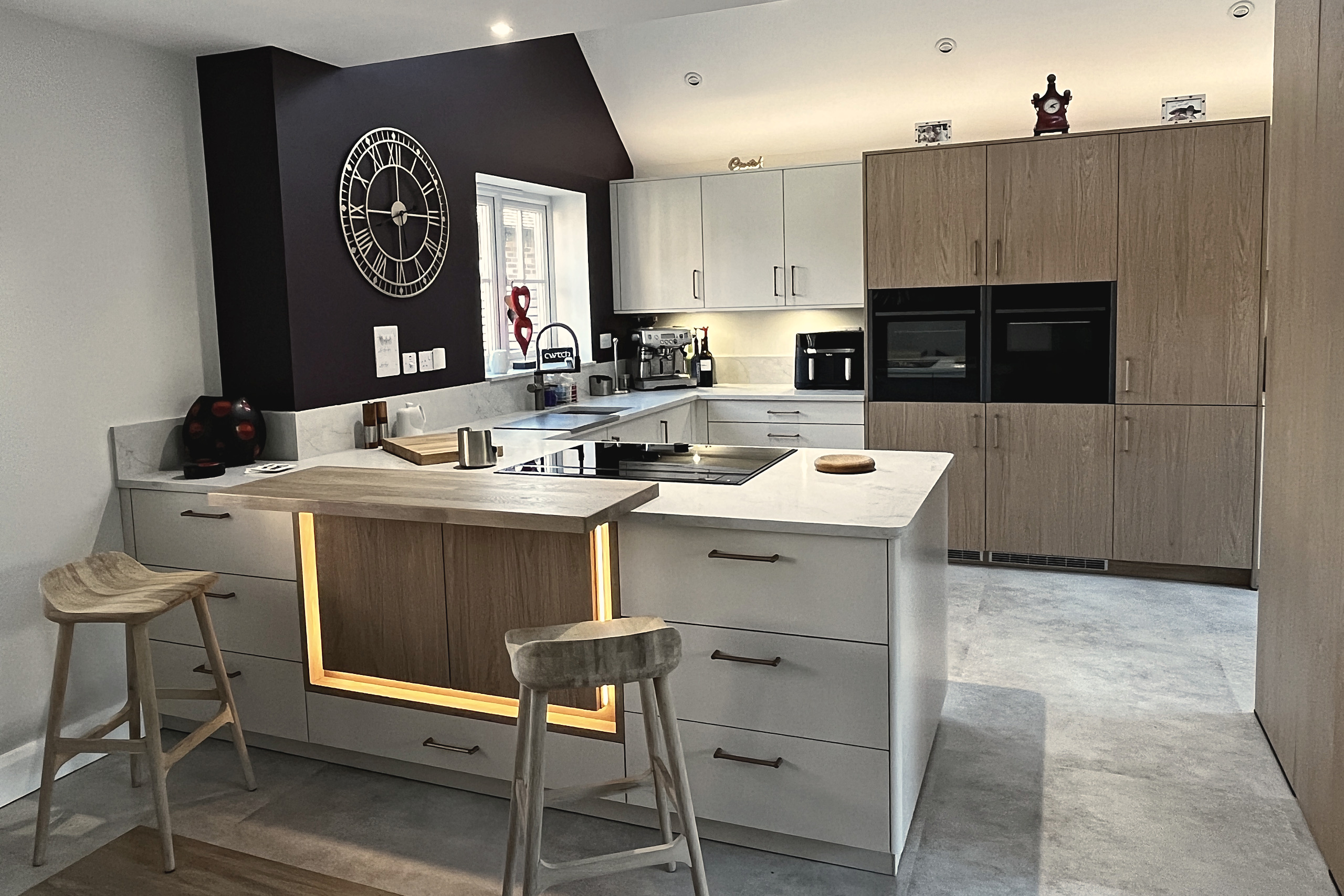 Pale grey kitchen with oak tall units, double ovens, white quartz worktops and a dark plum feature wall
