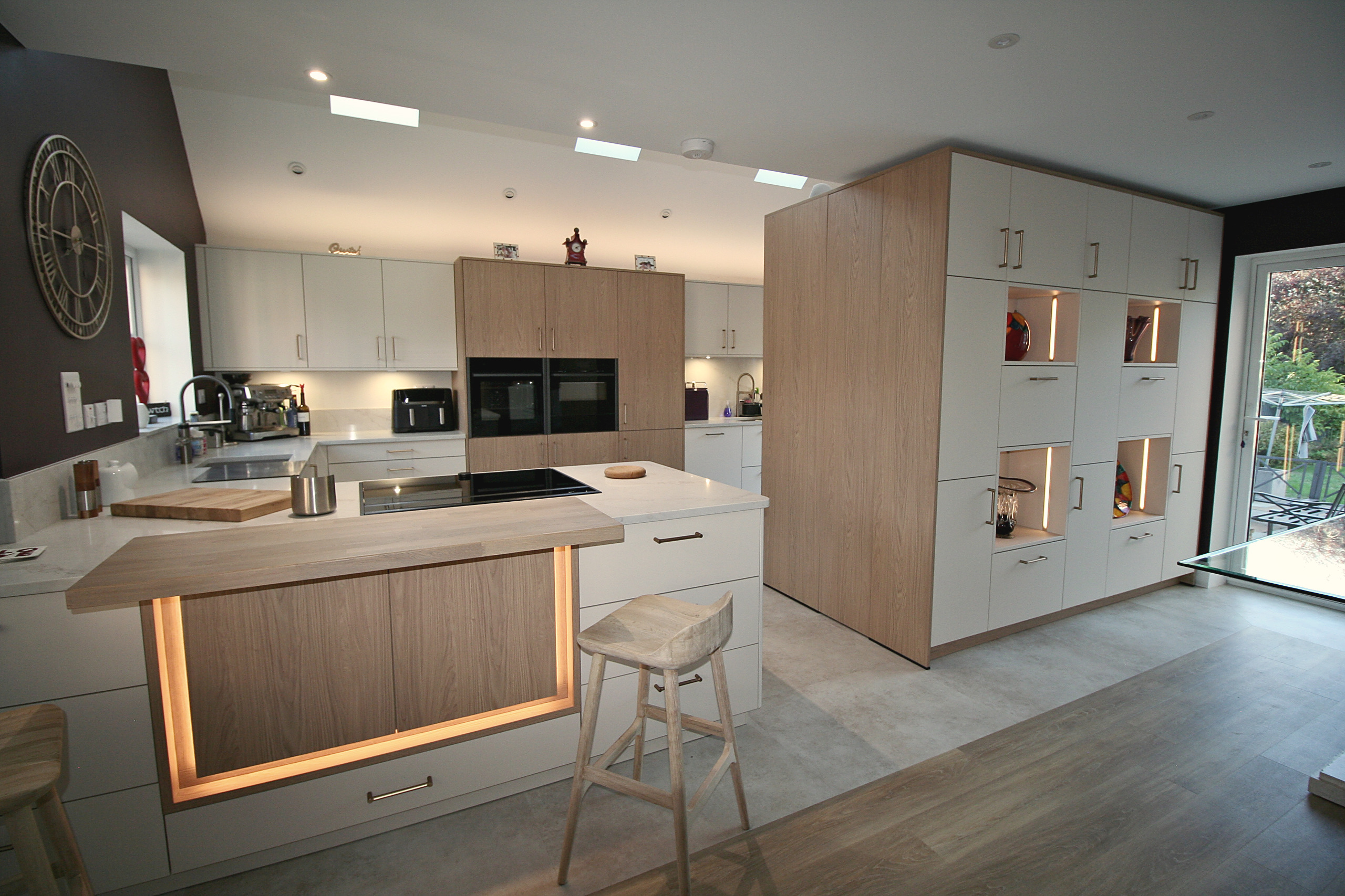 Cashmere and oak kitchen with illuminated breakfast bar, oak stool and tall lit display cabinetry