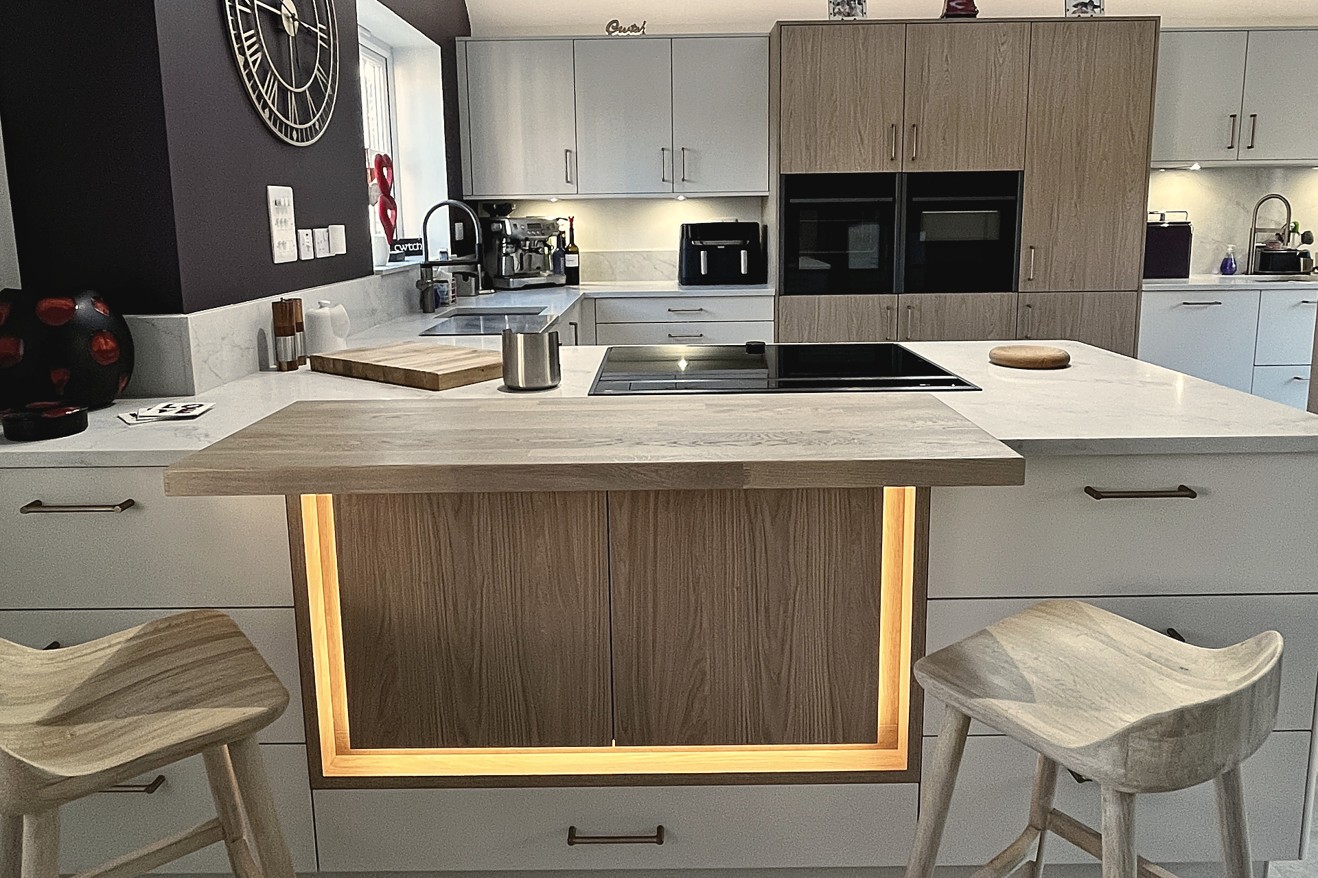 Island with oak breakfast bar, warm strip lighting, two timber stools and white quartz worktop