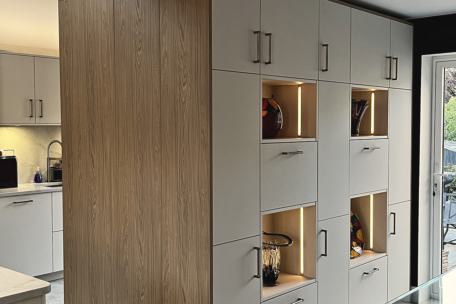 Oak and pale grey tall cabinet run with warm-lit open display niches beside a glazed garden door