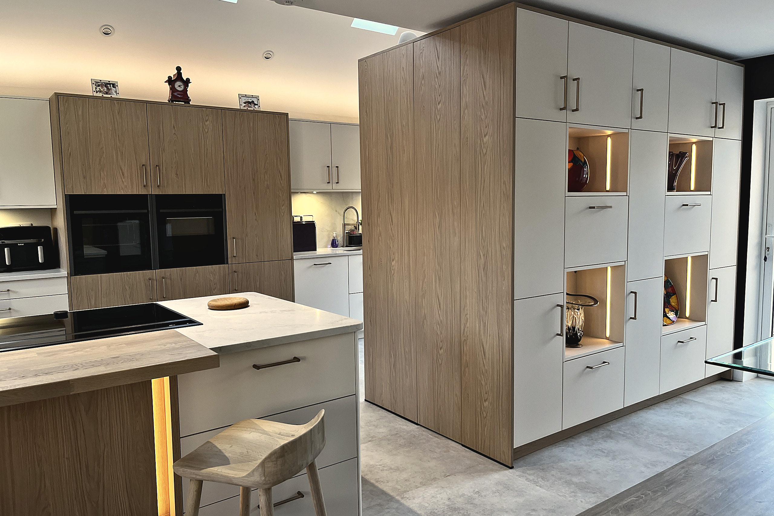 Kitchen island with quartz top and lit plinth beside a tall oak and cashmere wall with lit niches