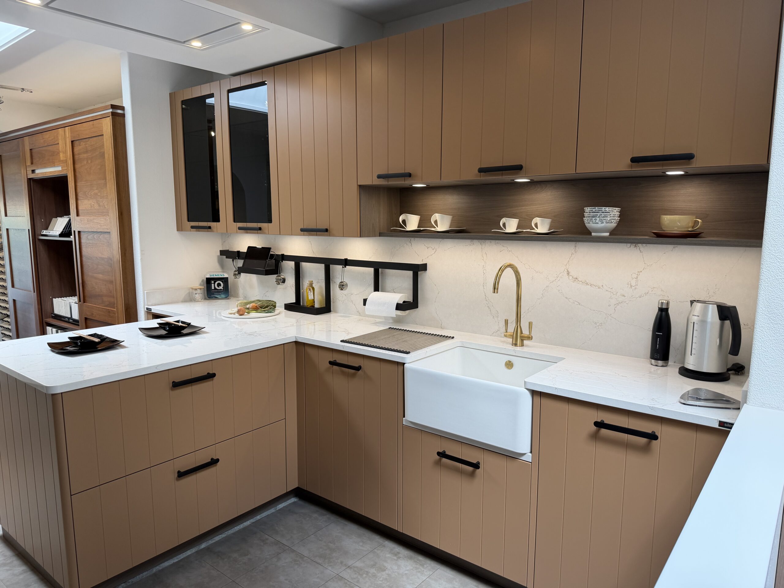 A showroom kitchen run in warm taupe beaded doors, with a white belfast sink and a brushed brass tap