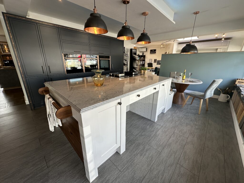 A wide view of the Kitchensmiths showroom: a marble-topped island under copper pendants, with a dining setting beyond