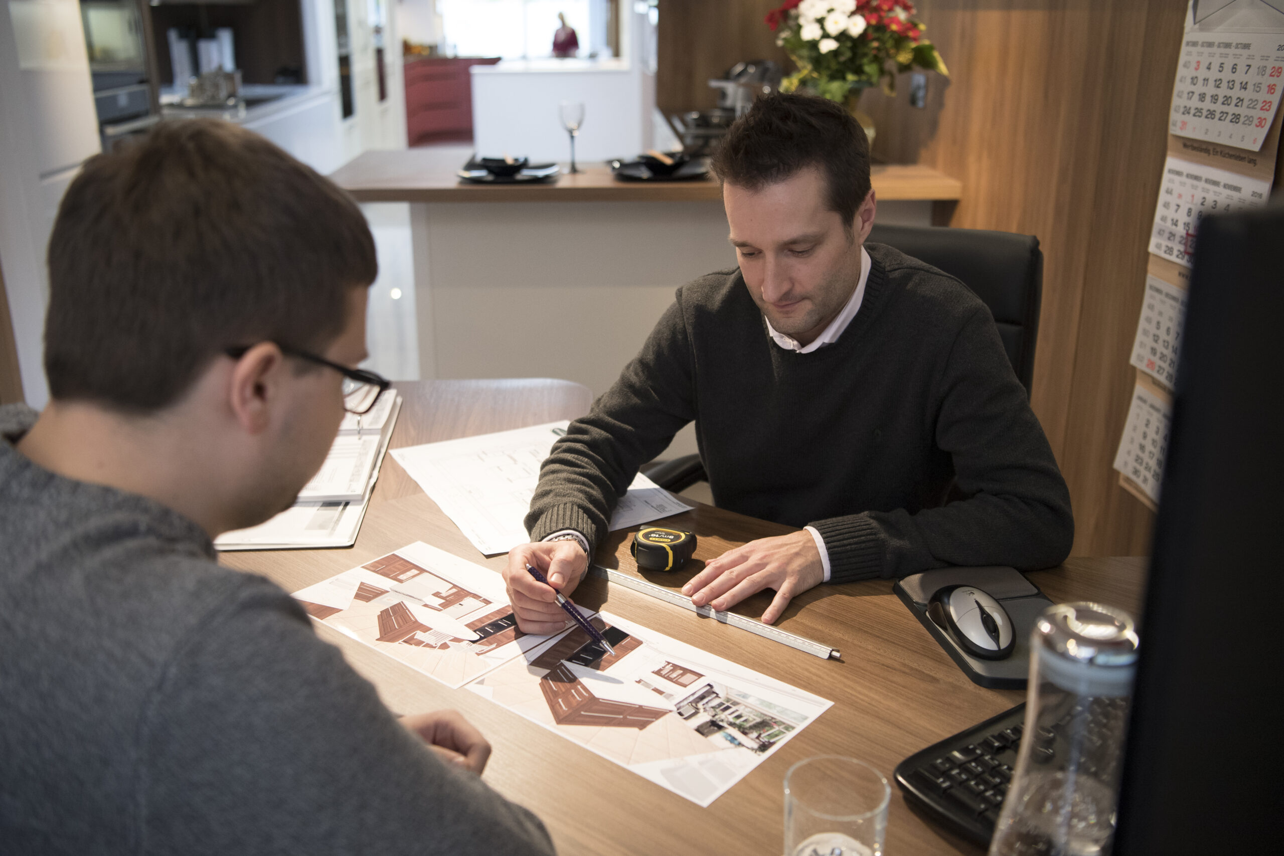 A Kitchensmiths designer and a client at a desk with printed plans, elevations, a scale rule and a tape measure