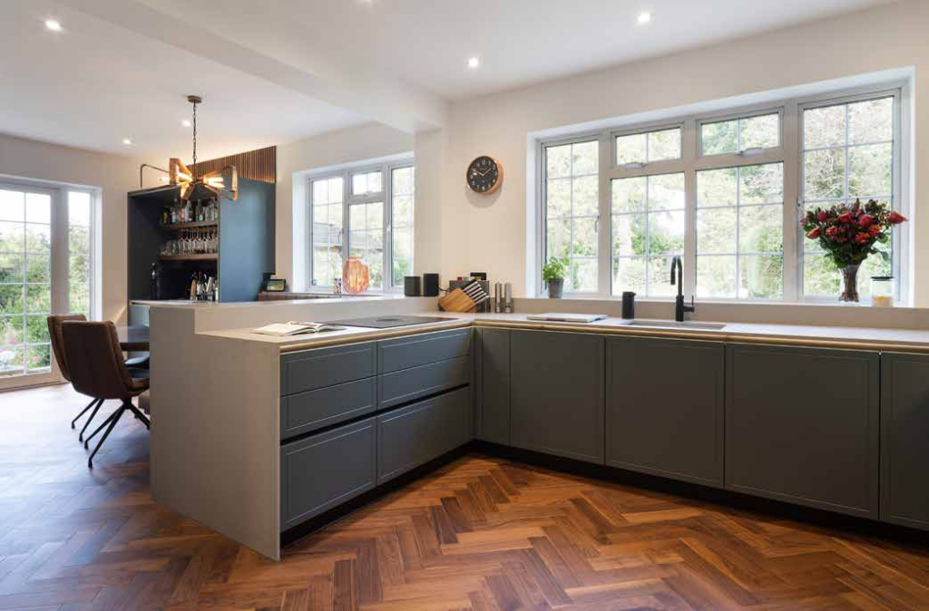 A grey kitchen whose run returns as a peninsula, dividing the working kitchen from a dining area with a herringbone floor.
