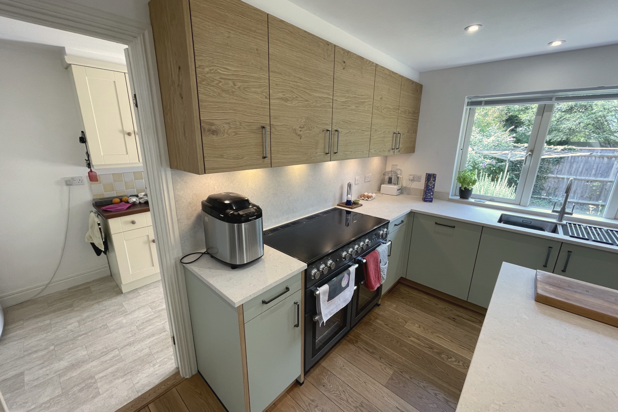 Range cooker and sink run in sage green with oak wall units and a window over the worktop