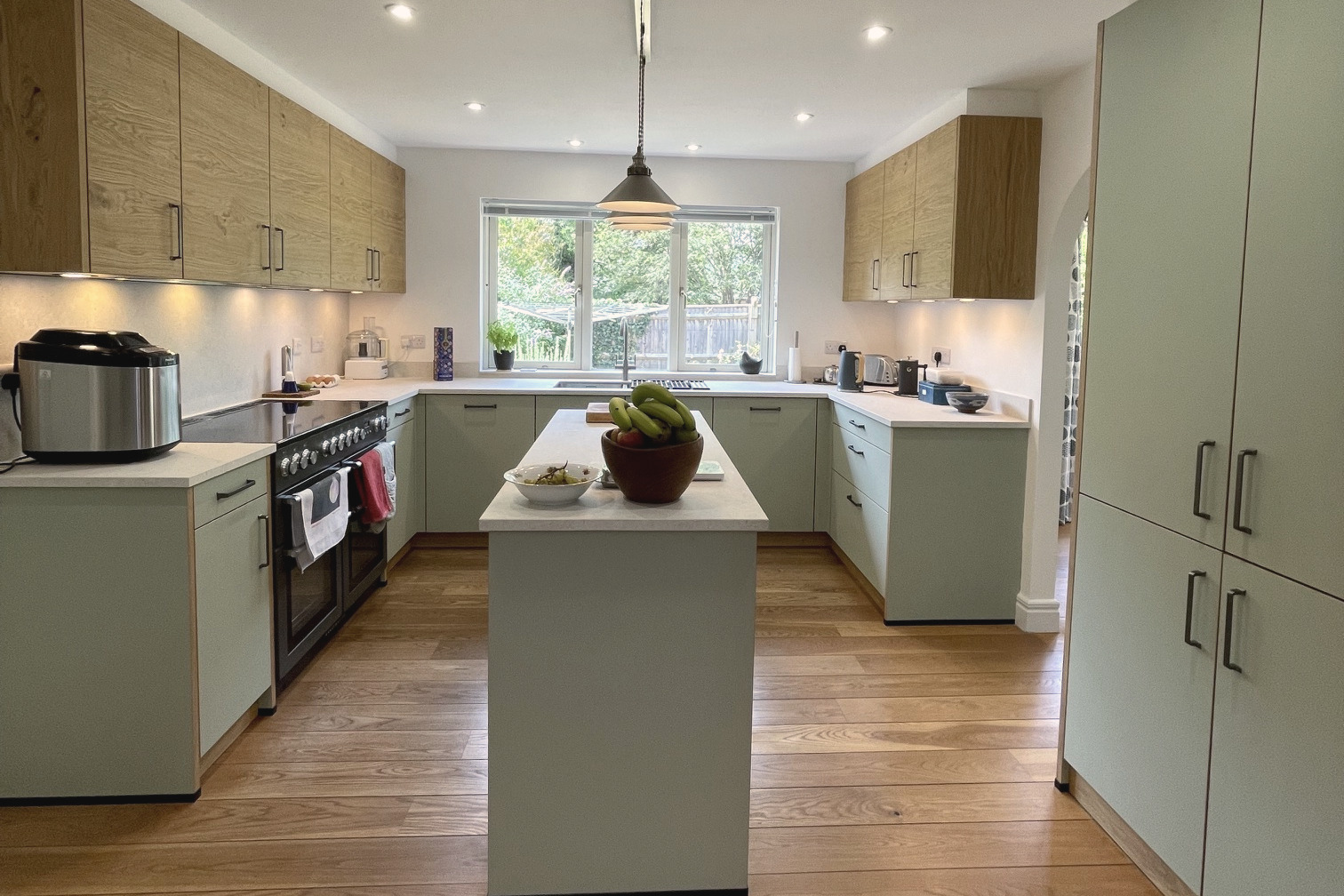 Symmetrical view of a sage green kitchen with oak wall units, central island and single glass pendant